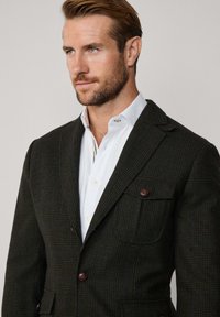Mörk rutig blazer med bruna knappar, två framfickor och notch-lapel, som bärs med en vit skjorta under.