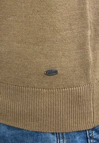Brauner Strickpullover mit geripptem Saum, versehen mit einem kleinen Metallschild mit "Peirrot"-Markierung. Die Textur wirkt weich und mittleres Gewicht.