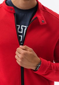 Giacca rossa con zip realizzata in tessuto morbido, con colletto alto, zip nera e una maglietta blu navy a contrasto sotto.