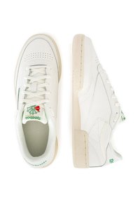 Baltos sportinės bateliai, pagaminti iš odos ir sintetinių medžiagų, su perforuotomis detalėmis, raištelių užsegimu ir tekstūriniu guminiu padu. Žalias logotipo akcentas.