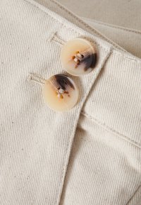 Deux boutons ronds dans des teintes beige et marron avec une finition brillante, comportant quatre trous, cousus sur un tissu texturé de couleur claire.