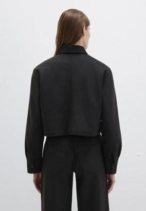 Woman with long brown hair wearing a black cropped jacket and high-waisted black pants, facing a plain white wall.