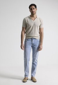 Camisa polo beige de manga corta con cuello en V, combinada con jeans ajustados azul claro y mocasines marrones, de pie contra un fondo neutro.