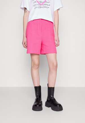 Person trägt knallpinke, strukturierte Shorts, weißes Grafik-T-Shirt und schwarze klobige Plateaustiefel, steht vor einem einfachen hellen Hintergrund.