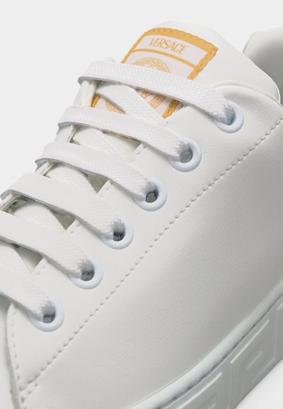Baskets blanches Versace avec lacets blancs, écusson logo doré sur la languette et bord de semelle en caoutchouc texturé.