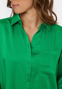 Green button-up shirt made of glossy fabric, featuring a collar, side pocket, and matching buttons. The texture is smooth and shiny.