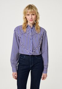 Blonde vrouw in een blauw-witte verticaal gestreepte blouse met een gerimpeld hartontwerp aan de voorkant, gecombineerd met donkerblauwe jeans en gouden grote oorbellen.