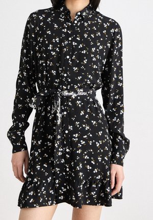 Kvinde iført en sort blomsterknap-kjole med lange ærmer og et bundet bælte i taljen med teksten "Tommy Jeans".