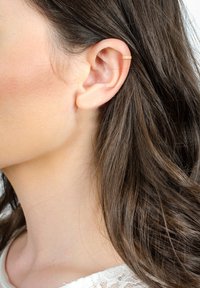 Guldörring prydd med intrikata utskärningar, bärs på den övre delen av örat. Långt, vågigt brunt hår flödar ner längs sidan.