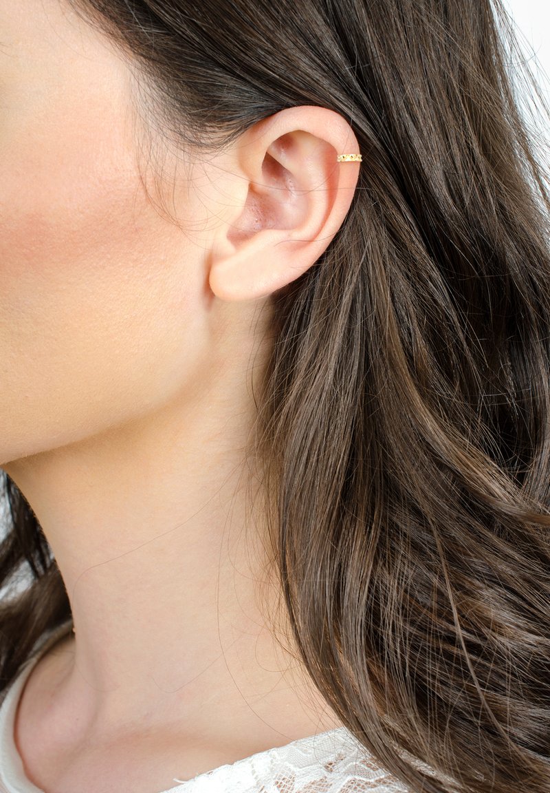 Guldörring prydd med intrikata utskärningar, bärs på den övre delen av örat. Långt, vågigt brunt hår flödar ner längs sidan.