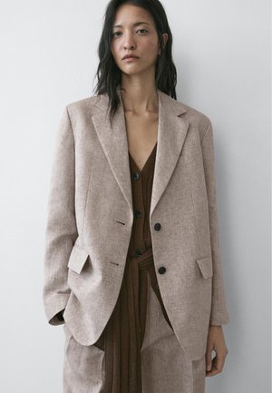 Junge Frau mit langen dunklen Haaren trägt einen beige oversized Blazer über einem braunen knöpfbaren Strickkleid und steht vor einer schlichten hellen Wand.