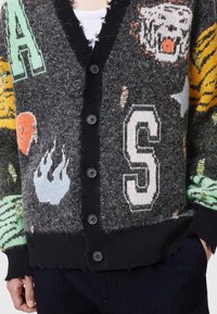 Pletený cardigan v šedých a černých barvách, s výraznými grafikami, žebrovanými knoflíky, viditelnými ošoupanými okraji a zvířecími motivy v jasných odstínech.