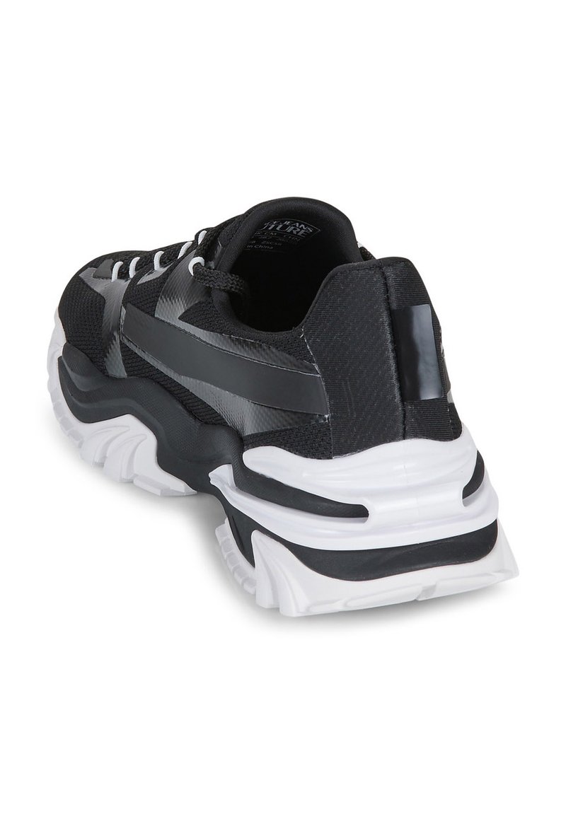 Zapatilla deportiva en blanco y negro con una parte superior texturizada, diseño elegante, suela gruesa y acentos reflectantes en un tacón voluminoso.