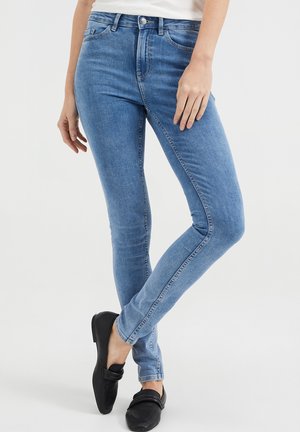 Femme portant un jean skinny bleu clair et des mocassins noirs, debout jambes croisées sur un fond uni.