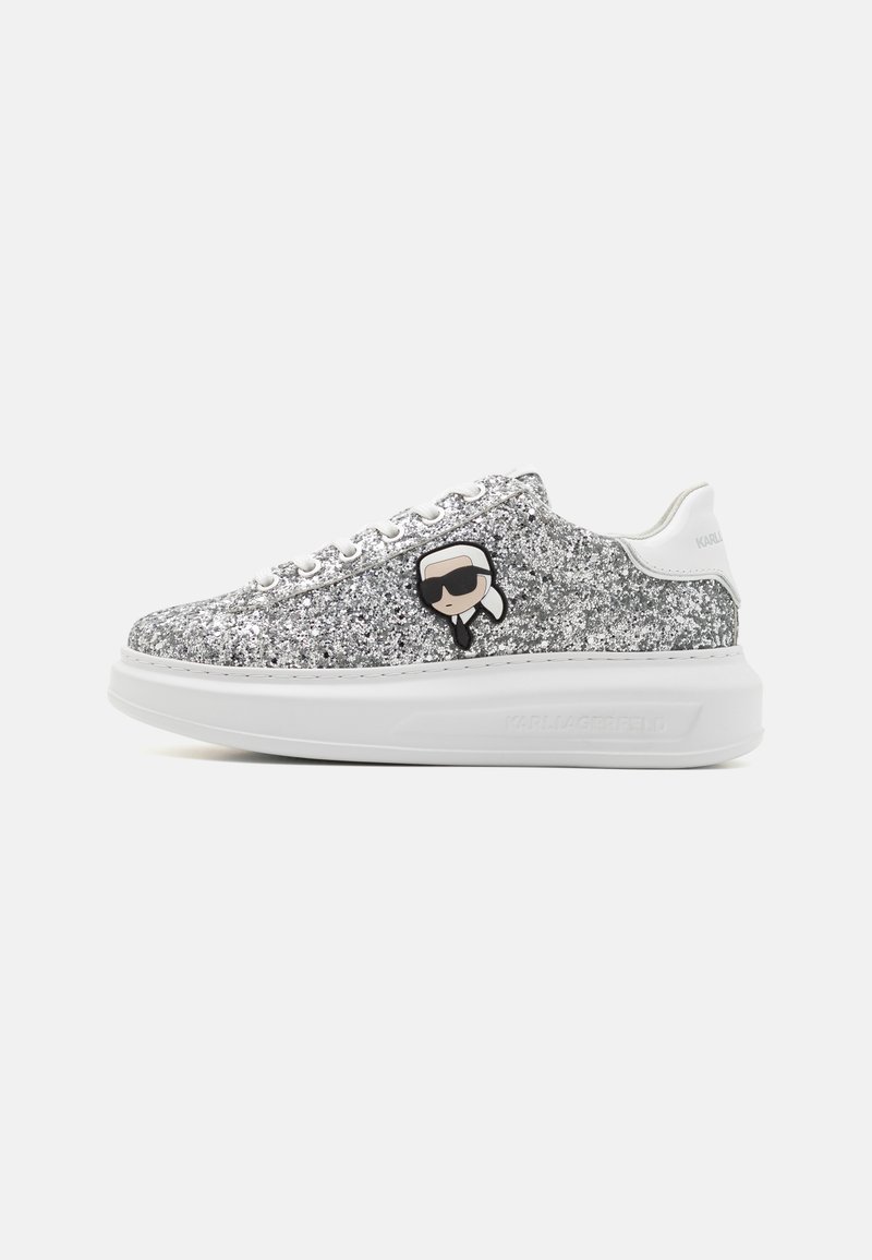 Zapatilla plateada brillante con una suela blanca gruesa, que presenta un logo de caricatura negro en el costado y detalles metálicos en los ojales.