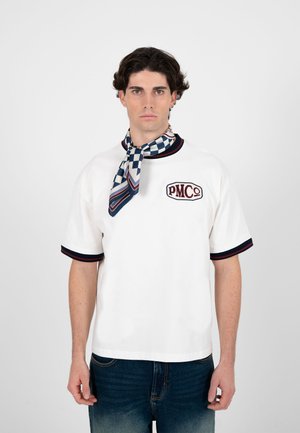 Ung mand med mørkt hår iført en hvid kortærmet skjorte med PMC-logo og ternet tørklæde bundet om halsen, stående foran en ensfarvet baggrund.