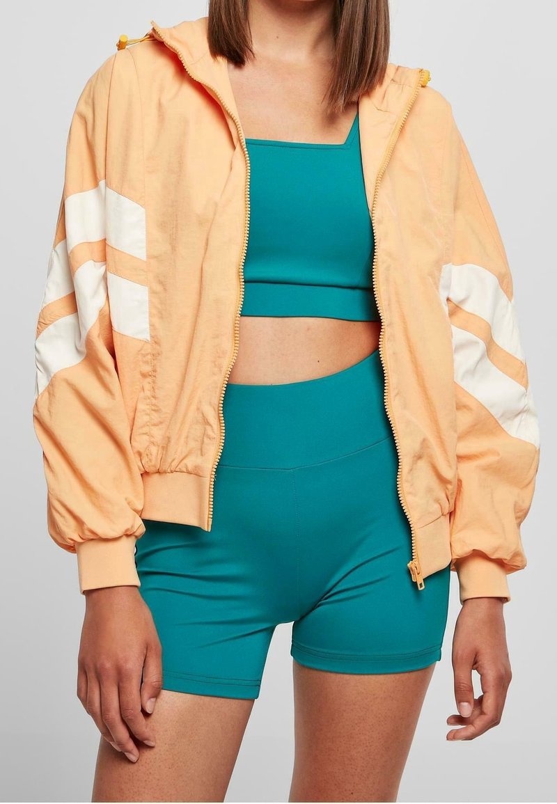 Femme portant une veste zippée pêche avec des rayures blanches, un soutien-gorge de sport teal et un short taille haute, debout avec les bras détendus.