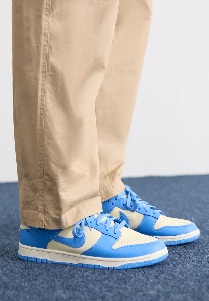 Zapatillas bajas en azul y crema con parte superior de cuero suave, puntera perforada y suela de goma texturizada, combinadas con pantalones de algodón beige.