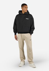 Black hooded sweatshirt with front pocket, and beige ribbed pants featuring a small logo. Model wearing white sneakers. Simple, casual design.