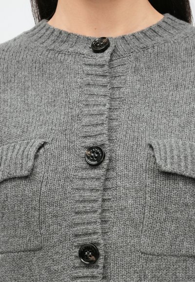 Κοντινό πλάνο γκρι πλεκτής ζακέτας με μαύρα κουμπιά και δύο τσεπάκια με καπάκια στο στήθος.