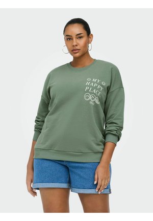 Nő visel olívazöld pulóvert, amelyen a "MY HAPPY PLACE" felirat és mosolygós arcok láthatók, hozzá felhajtott farmer rövidnadrág és karika fülbevalók.