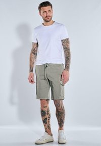 Khaki cargo shorts with side pockets, paired with a fitted white T-shirt and white sneakers. Skin visible, adorned with various tattoos.