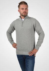 Grauer Reißverschluss-Pullover mit strukturiertem, geripptem Muster, langen Ärmeln und hohem Kragen. Getragen mit blauen Jeans.