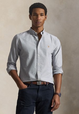 Mann mit hochgekrempelten Ärmeln, trägt ein hellblaues Hemd mit Knöpfen, dunkle Jeans, braunen Gürtel und Armbanduhr, steht mit einer Hand in der Tasche.