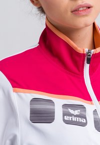 Weißer Sportjacke mit einem rosa und orangen Reißverschlusskragen. Das Design umfasst gestreifte Akzente und ein Logo auf der linken Seite.