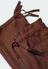 Pantalon en lin marron avec taille élastique et ceinture à nouer en tissu, avec des coutures visibles et des détails d’une coupe décontractée.