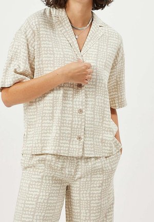 Kobieta ubrana w beżową koszulę na krótki rękaw z guziczkami i pasujące spodnie z abstrakcyjnym białym wzorem, ozdobiona warstwowymi srebrnymi naszyjnikami.