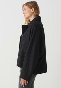 Giacca nera in misto lana con colletto alto, cinque bottoni, una tasca e maniche oversize, con una finitura testurizzata e una silhouette rilassata.