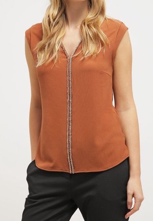 Vrouw draagt een mouwloze roestkleurige blouse met een V-hals en decoratieve verticale bies, gecombineerd met een zwarte broek.