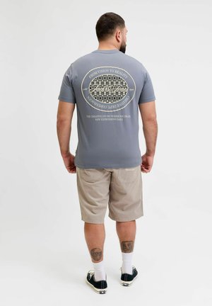 Mand med skæg iført grå grafisk t-shirt og beige shorts, stående med ryggen mod kameraet mod en ensfarvet hvid baggrund.