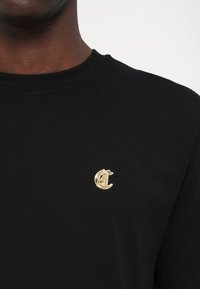Tricou din bumbac negru, cu un logo brodat aurie pe piept. Design cu guler rotund, mâneci scurte și o textură netedă.
