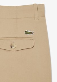 Pantaloni in cotone beige con tasca a patta, bottone tondo e logo coccodrillo ricamato verde sul lato.
