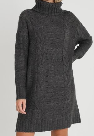 Robe-pull à col roulé en maille tricotée gris foncé avec des motifs torsadés et des manches longues, portée par une personne avec une main sur la hanche.
