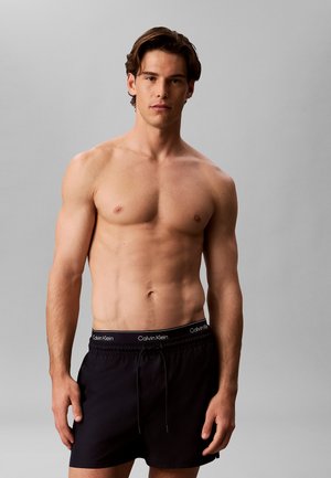 Mężczyzna bez koszulki z krótkimi brązowymi włosami, ubrany w czarne kąpielówki Calvin Klein na jednolitym szarym tle, zwrócony przodem.