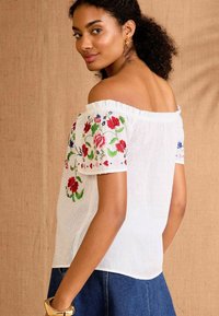 White off-shoulder blouse with floral embroidery in red, blue, and green on short puff sleeves. Textured fabric with a subtle stripe pattern.