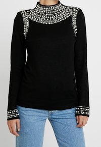 Svart långärmad topp med hög krage, prydd med klungor av vita pärlor på kragen, axlarna och ärmslut, bärs med ljusblå jeans.