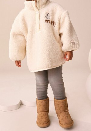 Enfant portant un pull polaire crème avec un logo de lapin, des leggings gris et des bottes beiges ornées de visages de lapin, debout sur un fond neutre.