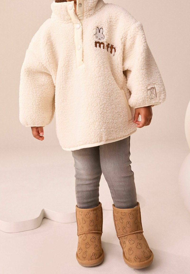 Enfant portant un pull polaire crème avec un logo de lapin, des leggings gris et des bottes beiges ornées de visages de lapin, debout sur un fond neutre.