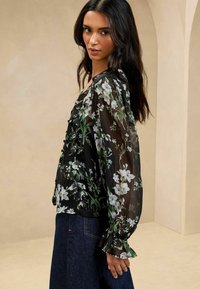 Schwarze transparente Bluse mit floralem Muster, langen Ärmeln, Rüschenausschnitten und Knopfdetails. Zu dunklen Jeans getragen.