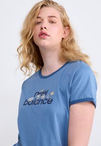 Mėlynas medvilninis marškinėliai trumpomis rankovėmis, su gėlių grafiniu dizainu ir tamsiomis raidėmis užrašas „new balance“, apvaliu kaklu.