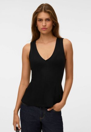 Young woman with long hair wears a sleeveless black V-neck top and dark blue jeans, standing with one hand in pocket and neutral expression.