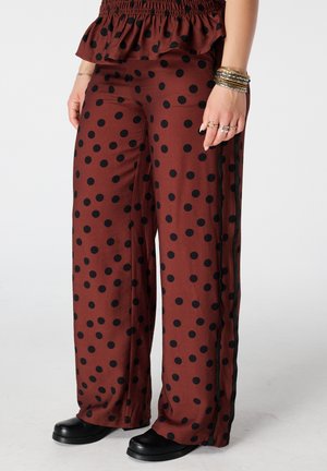Pantalon marron à jambes larges avec des pois noirs porté avec des chaussures noires, visible de la taille jusqu’aux pieds, accessoirisé de bracelets et de bagues.