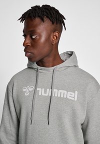 Grå hoodie med en stor vit grafisk text som står "tunnel." Har en dragsko för huva och en mjuk textur. Närbild av bärarens profil från sidan.
