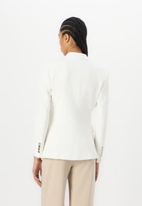 Hvit skreddersydd blazer med tett silhuett, spisse klaffer og sølvknappdetaljer på ermene; glatt tekstur og strukturert design.