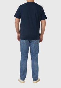 Homme debout, de dos, portant un t-shirt bleu marine, un jean bleu clair et des baskets blanches sur un fond gris.