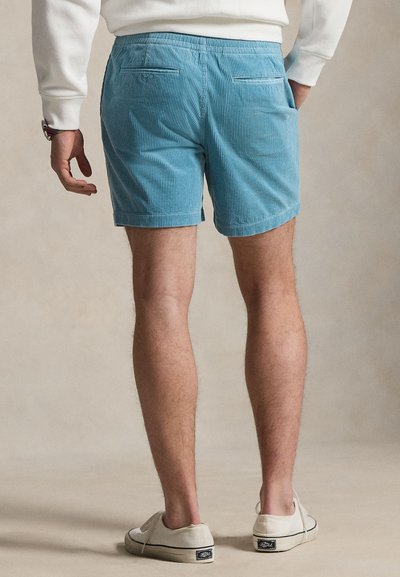 Άνδρας που φοράει ανοιχτό μπλε βερμούδα από κορδόνι, άσπρο φούτερ, άσπρα αθλητικά παπούτσια, στέκεται με το ένα χέρι στην τσέπη μπροστά σε ουδέτερο φόντο.